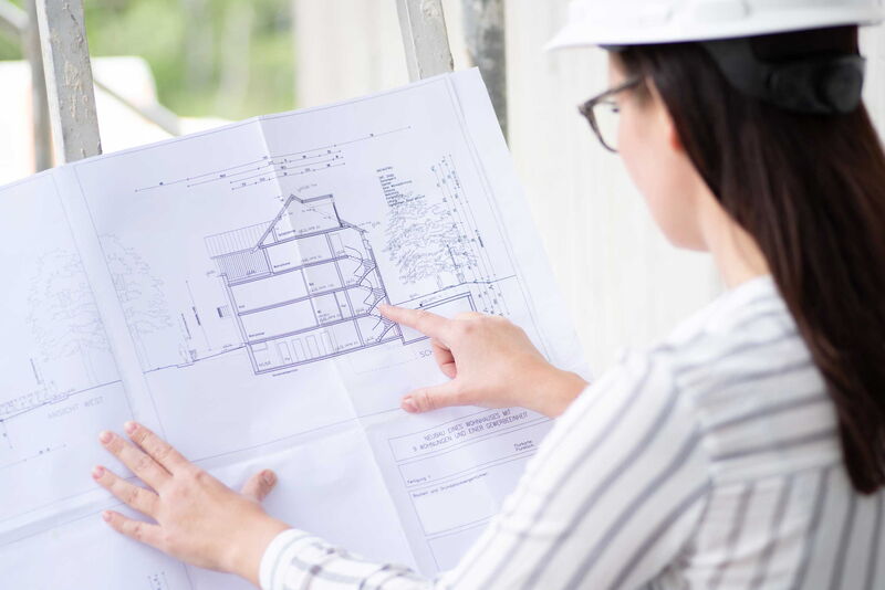 Weibliche Person mit Helm schaut und zeigt seitlich auf technische Zeichnungen.