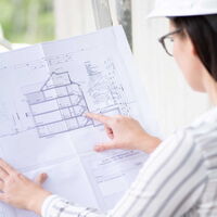 Weibliche Person mit Helm schaut und zeigt seitlich auf technische Zeichnungen.