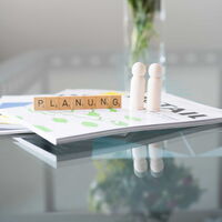 Aus Holzbausteinen wurde das Wort Planung gelegt, im Hintergrund links ist ein Blumenstrauss zu sehen.