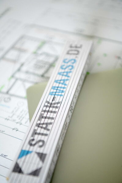 Ein Zollstock mit dem Logoaufdruck von Statik Maaß liegt auf Bauplänen.