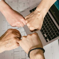 Vier Fäuste stoßen in der Mitte zusammen über einem Laptop und technischen Zeichnungen.