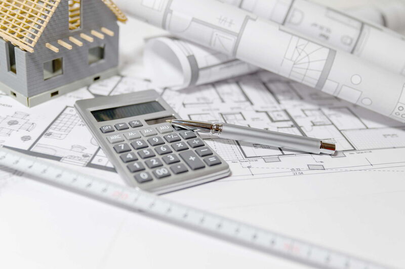 Oberfläche eines Tisches auf welchem Baupläne, ein Taschenrechner und ein Stift liegen, sowie ein kleines Modelhaus steht.